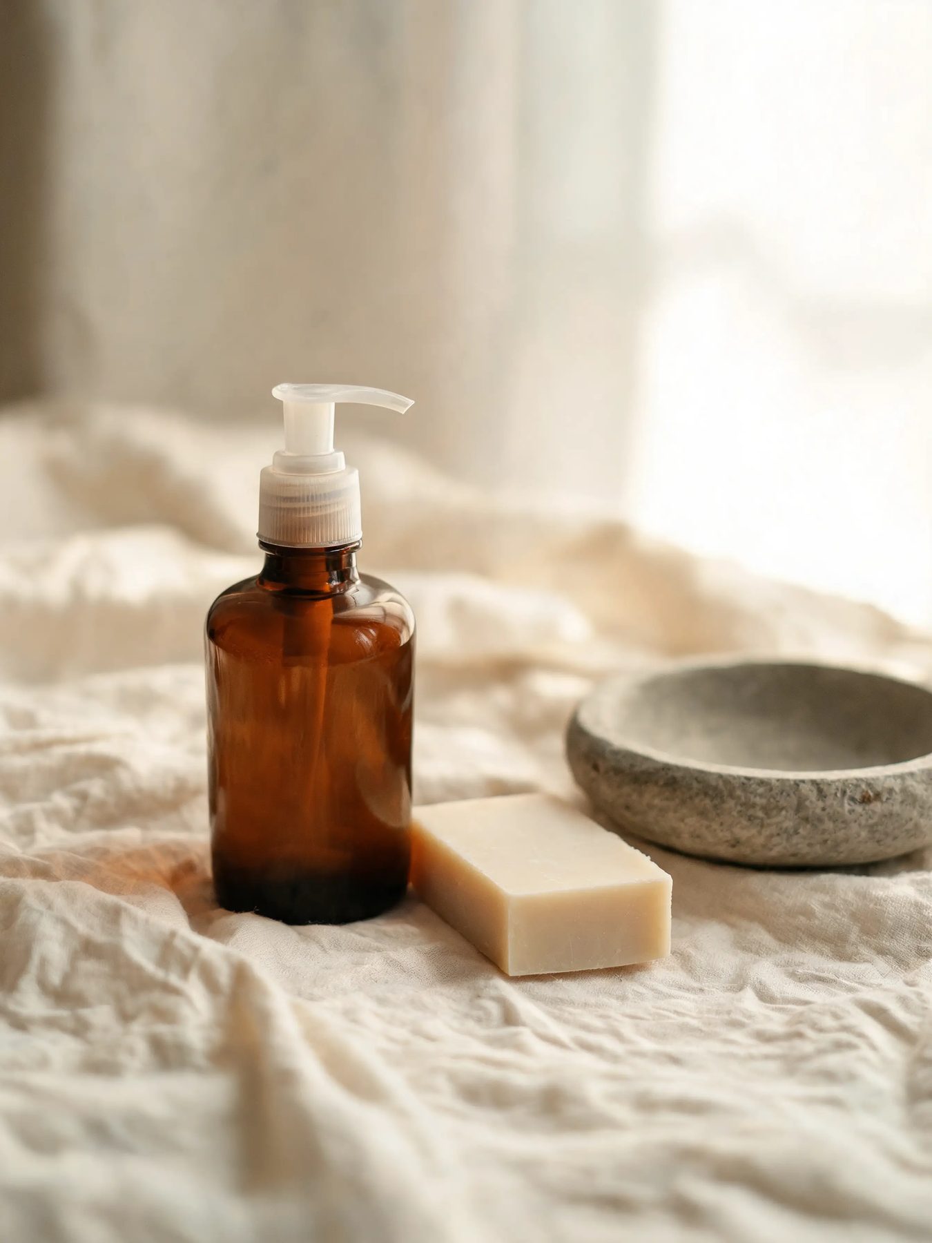 Amber glass bottle and a cut bar of soap on linen beside a stone basin